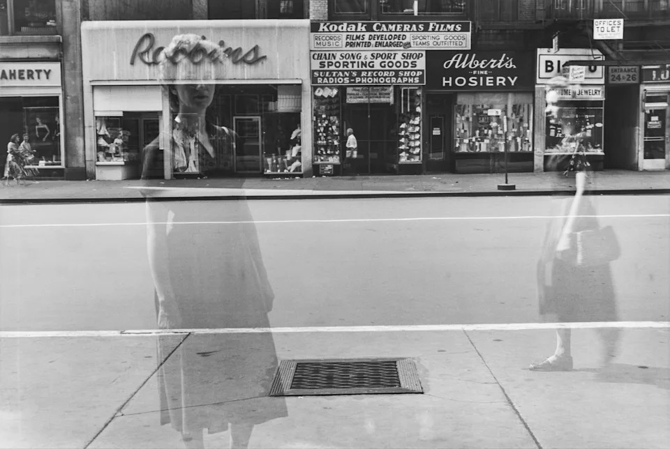 A double-exposure layers a ghostly female figure over a New York street scene, her translucent form standing in front of shop fronts including a Kodak Cameras Films sign.