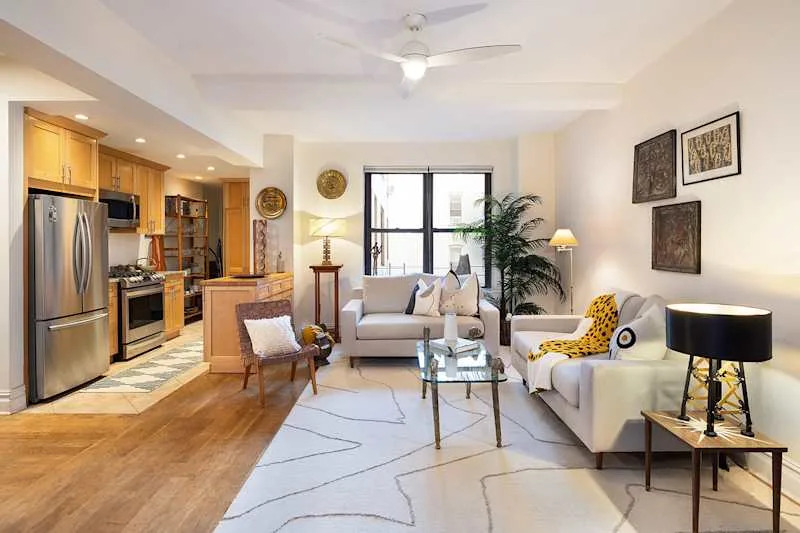 Bright living room with a light gray sofa, patterned rug, glass coffee table, and a kitchen area featuring wooden cabinets.