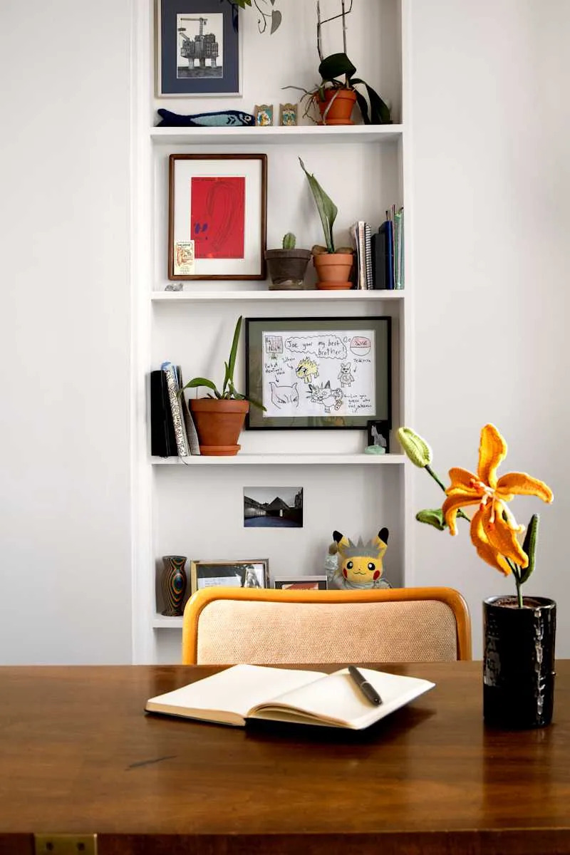 A wooden desk with an open notebook and pen, a black vase with a yellow flower, and a shelf filled with plants and framed art.