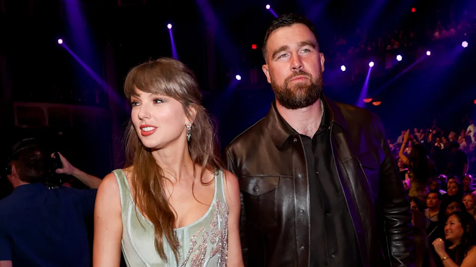 Taylor Swift, in a light dress, stands beside Travis Kelce in a leather jacket at an event with a crowd and purple lights.