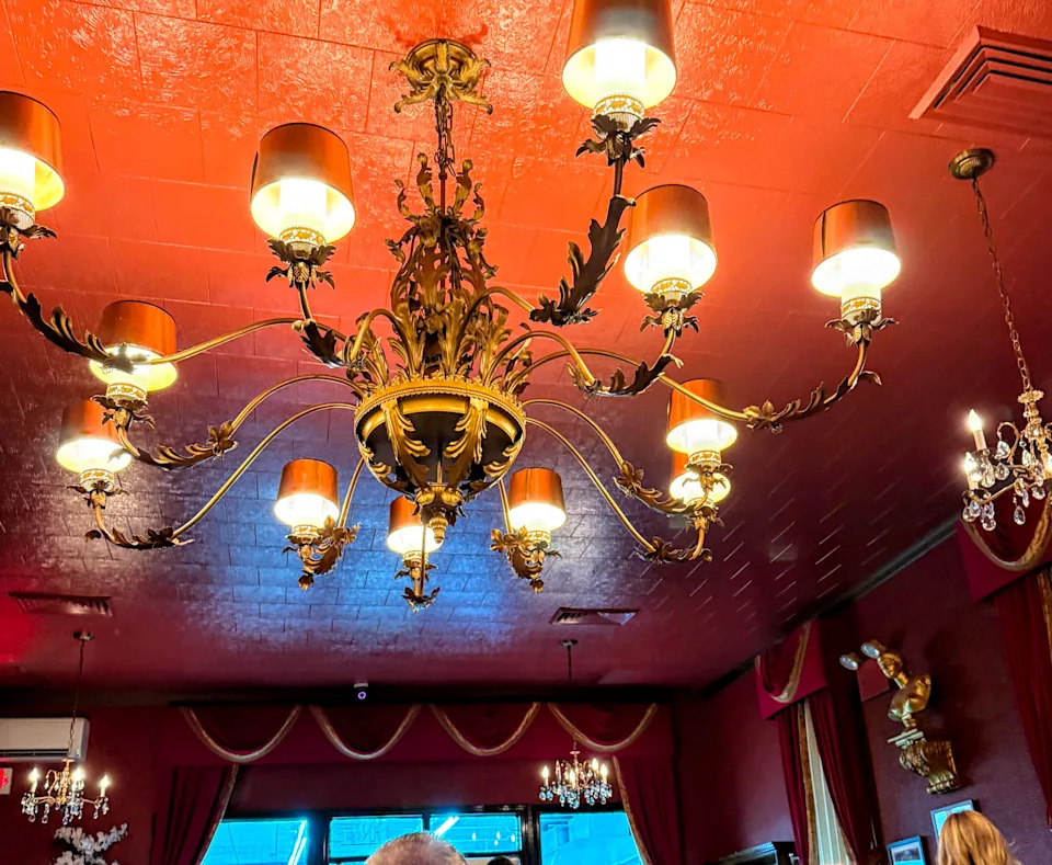 An elaborate gold-colored chandelier with individually-shaded light bulbs hangs from a red ceiling.