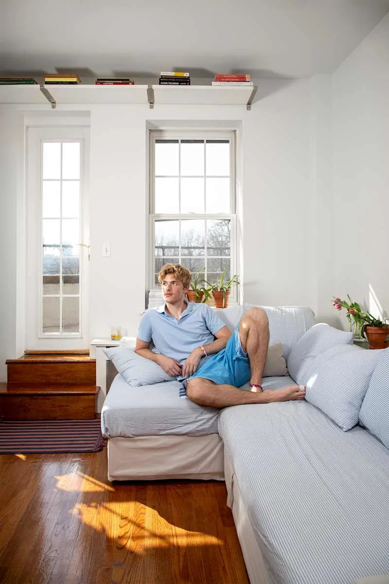 A young man in a light blue polo and shorts sits on a striped couch, with plants and bookshelves nearby.