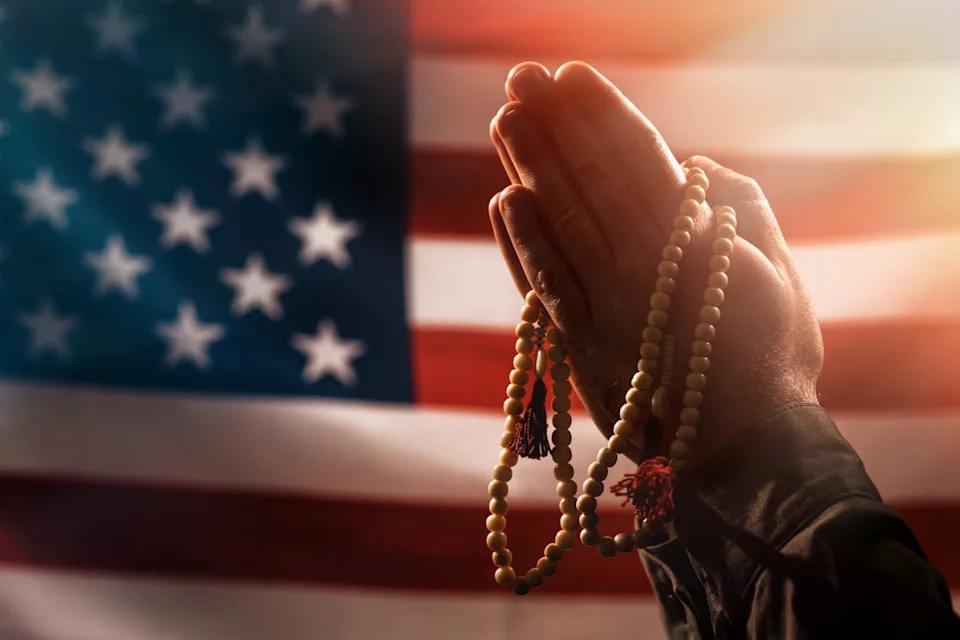 Male hands folded in prayer holding a rosary with an American flag in the background