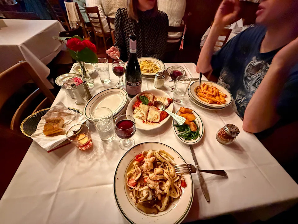 Three people are gathered around a white-clothed table, each with a white dish of pasta and a glass of red wine. A plate of red-sauce covered chicken, a plate of vegetables, a basket of bread, and a bottle of wine share the table.