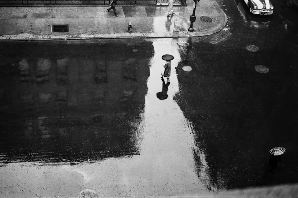 Looking down from above onto a rain-flooded Lexington Avenue, a single figure with an umbrella stands at the centre of a great pale reflection, tiny against the wet street.