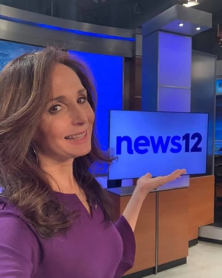 Former news anchor Amy McGorry in a news studio, gesturing towards a screen displaying "news12".