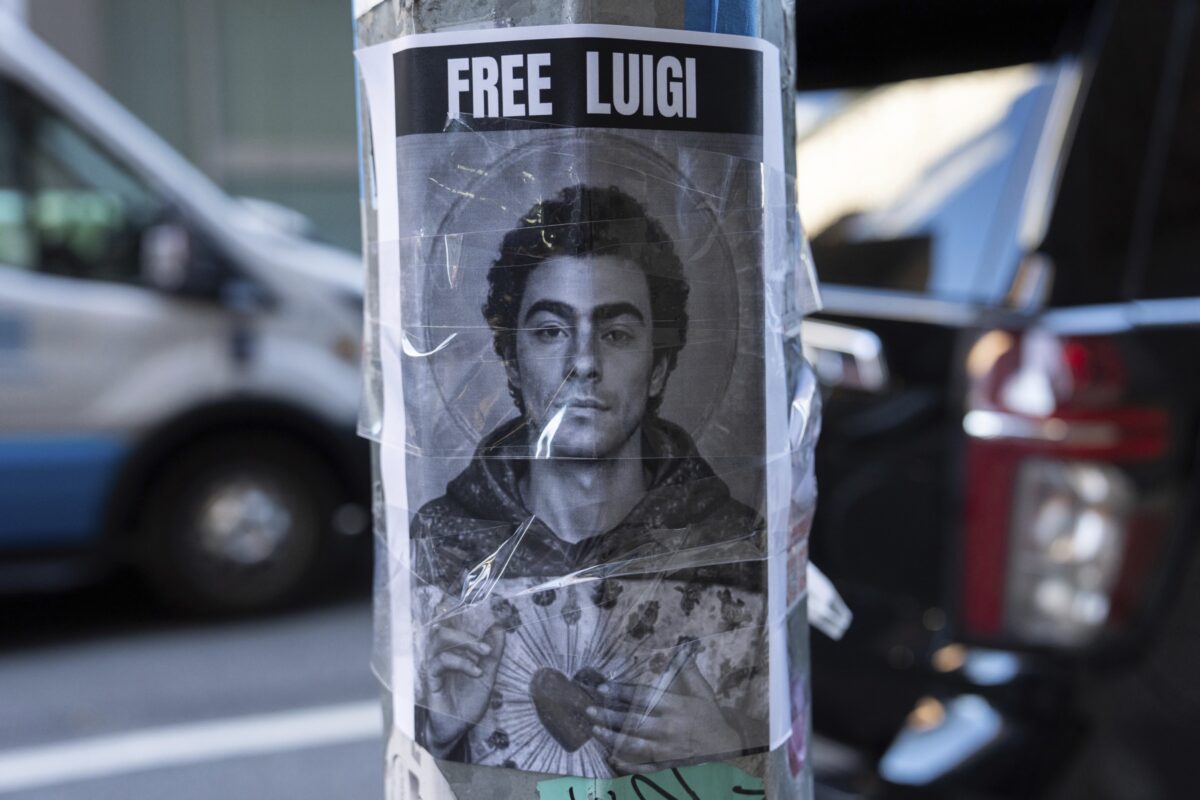 A black-and-white poster taped to a street pole shows a young man resembling a saint-like figure, with a halo-like design behind his head and his hands holding a symbolic heart. Bold text above reads 