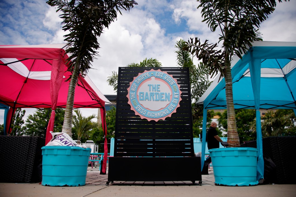 A general view of "The Garden" in Staten Island, New York, featuring a pink tent and a blue tent with palm trees in between them, and a sign for "The Garden, Est. 2026, 2655 Richmond Ave SI NY" in the background.