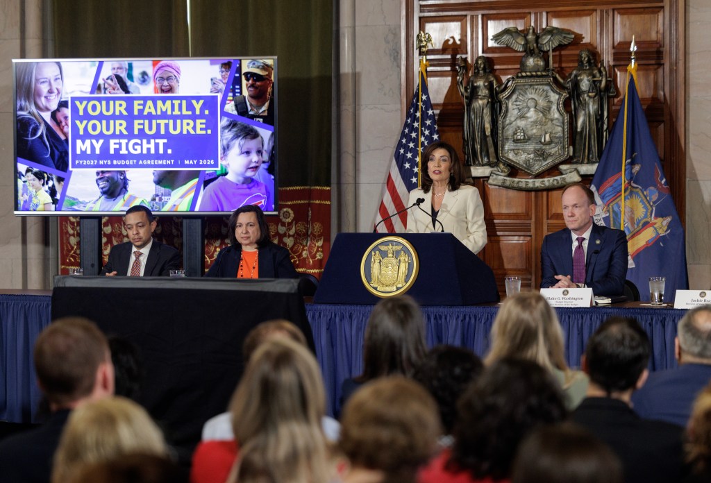 Governor Kathy Hochul making a budget announcement with a screen behind her displaying "YOUR FAMILY. YOUR FUTURE. MY FIGHT. FY2027 NYS BUDGET AGREEMENT | MAY 2026."