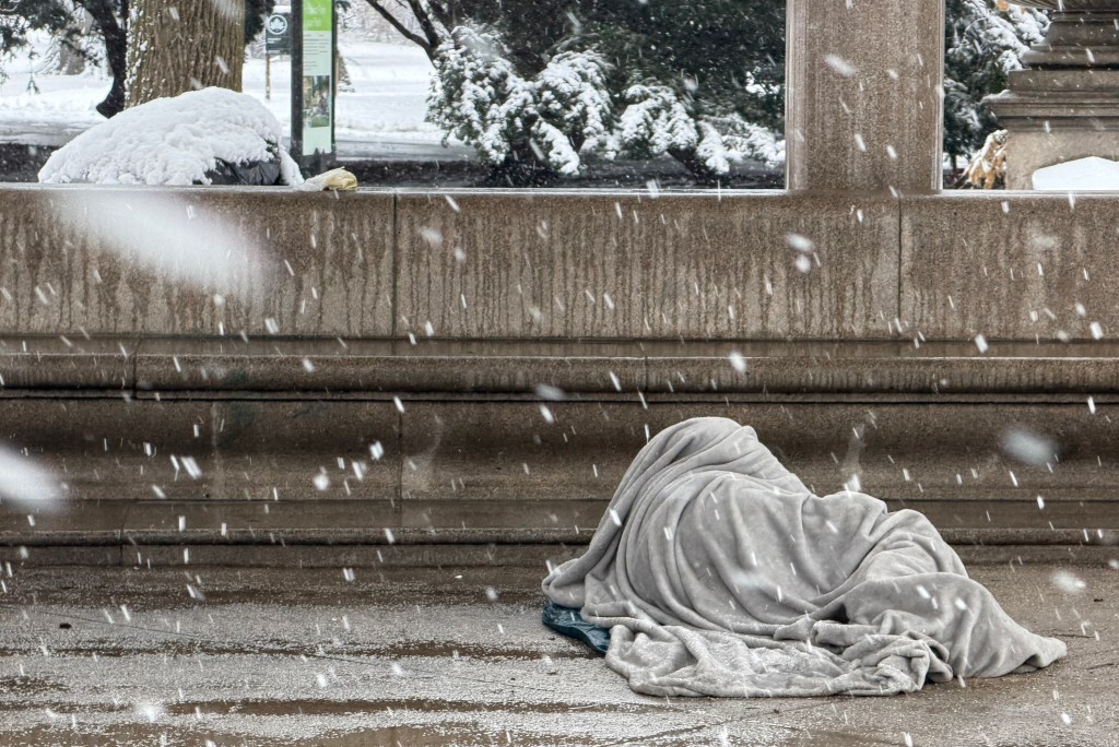 A homeless person sleeping under a blanket at the entrance of Prospect Park in Brooklyn during a snowstorm.