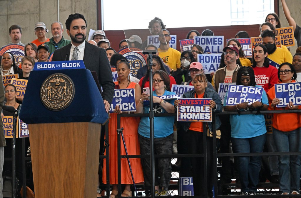 Mayor Zohran Mamdani speaking at a podium in front of a crowd holding signs that read "Block by Block" and "Invest in Stable Homes".