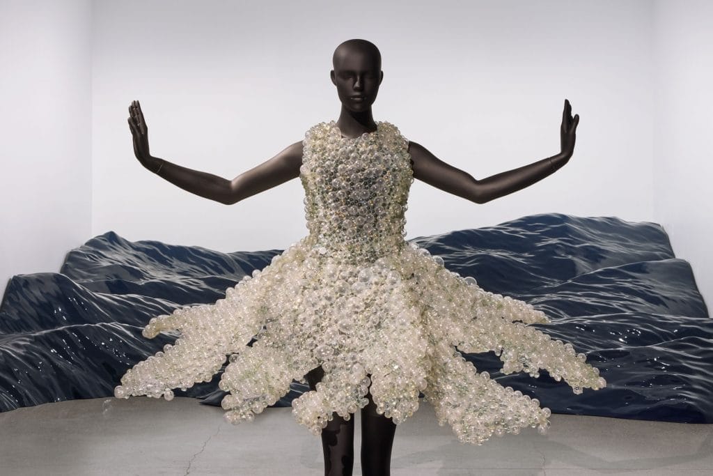 Mannequin wears shimmering bubble-like dress beside undulating blue installation, arms extended symmetrically outward dramatically.