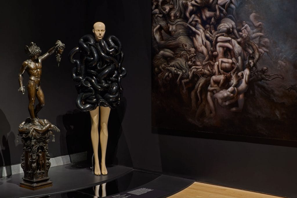 Black sculptural dress displayed beside classical statue and dark mythological painting inside atmospheric exhibition gallery.