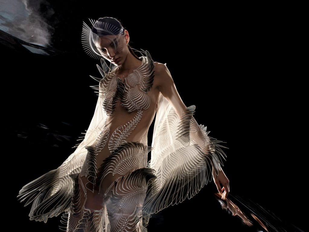 Model wears intricate translucent sculptural garment with radiating filaments and biomechanical details against dark background.