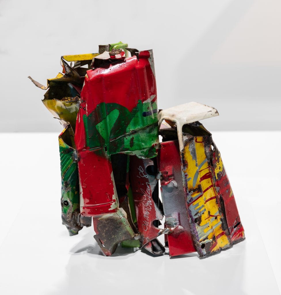 Abstract metal sculpture made from crushed and twisted sheet metal, painted in bright red, green, and yellow with areas of wear and rust. The jagged pieces are bent and layered into a compact, upright form, with sharp edges, dents, and overlapping panels suggesting repurposed car parts. The sculpture sits on a plain white surface against a neutral gray background. Featured at the 2026 edition of TEFAF New York.