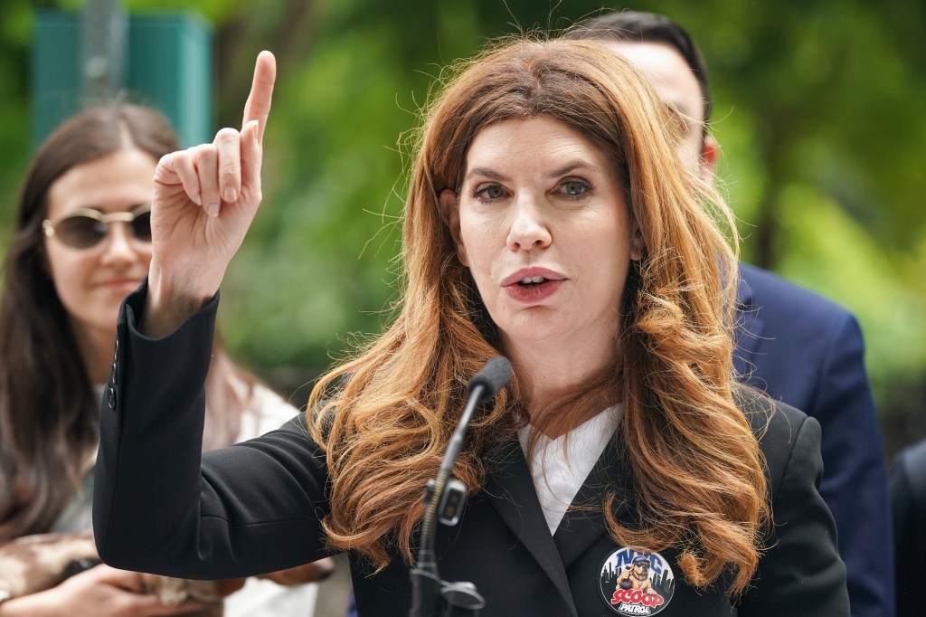 Julie Menin, Speaker of the NYC Council, speaks into a microphone at a press conference, with her right hand raised and index finger pointing upwards.