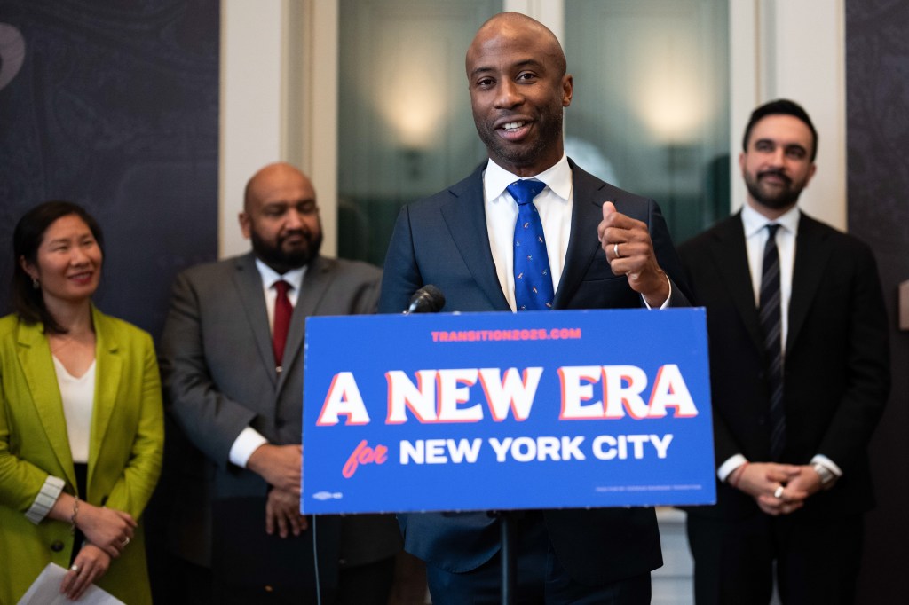Kamar Samuels speaking at a podium with "A NEW ERA for NEW YORK CITY" on it, with three people standing behind him.