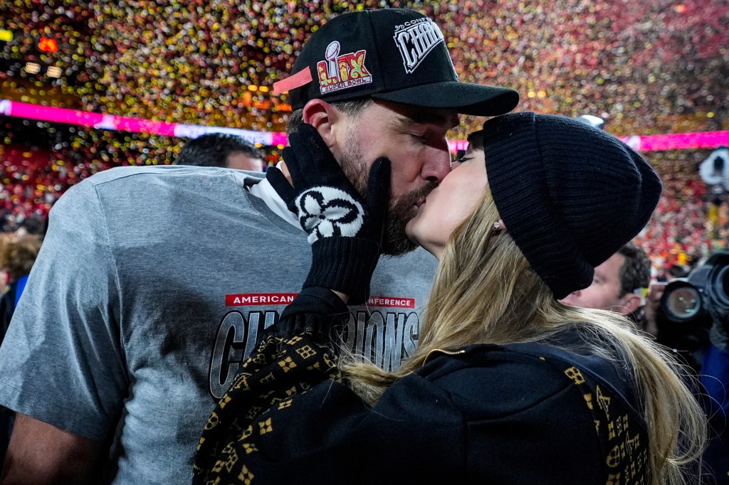 Travis Kelce wearing a Super Bowl LIX hat and Taylor Swift in a black beanie sharing a kiss.