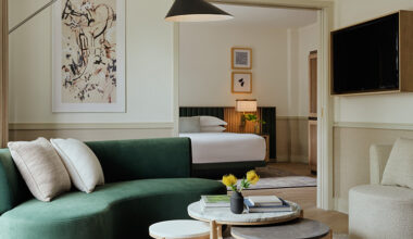 Modern hotel suite living area with a curved green sofa, layered round coffee tables, and an arched floor lamp, opening into a bedroom with a neatly made bed, abstract wall art, and soft neutral décor.
