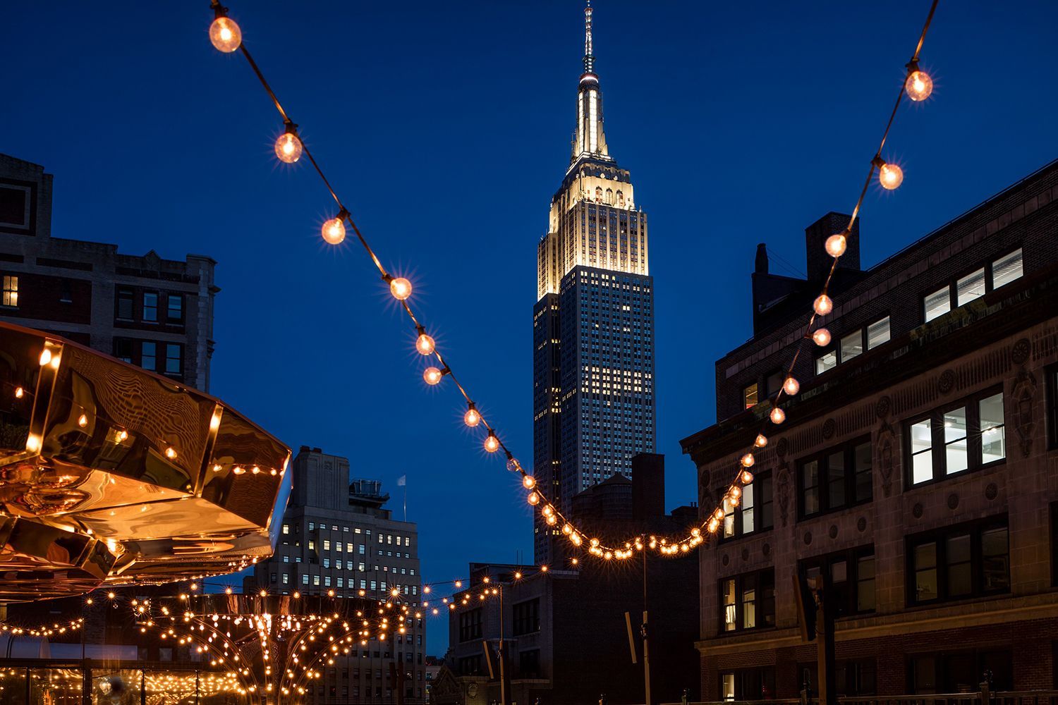 Evening view from the Magic Hour Rooftop Bar at Moxy Times Square, showcasing lounge seating and an unobstructed view of the illuminated Empire State Building.