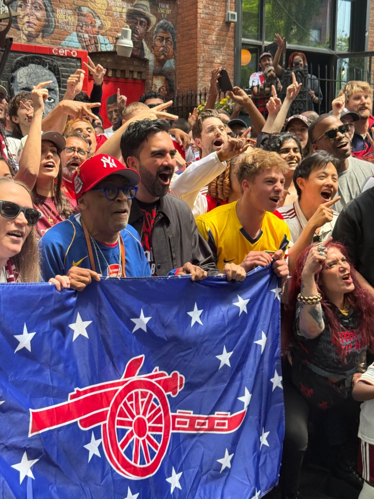 Mamdani and Spike Lee holding an Arsenal flag with fans at Fancy Free.