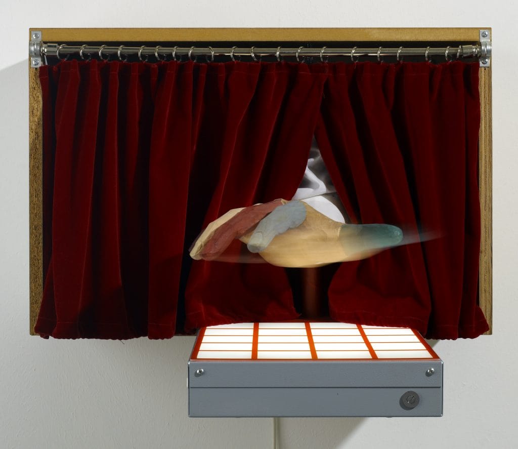 Wall-mounted installation featuring a small stage framed in wood with deep red curtains drawn mostly closed. From between the curtains, a carved, multicolored hand-like form extends outward, appearing blurred as if in motion. Below the stage is a rectangular light box with a glowing grid pattern in orange and white, attached to a gray metal housing. Featured at the 2026 edition of TEFAF New York.