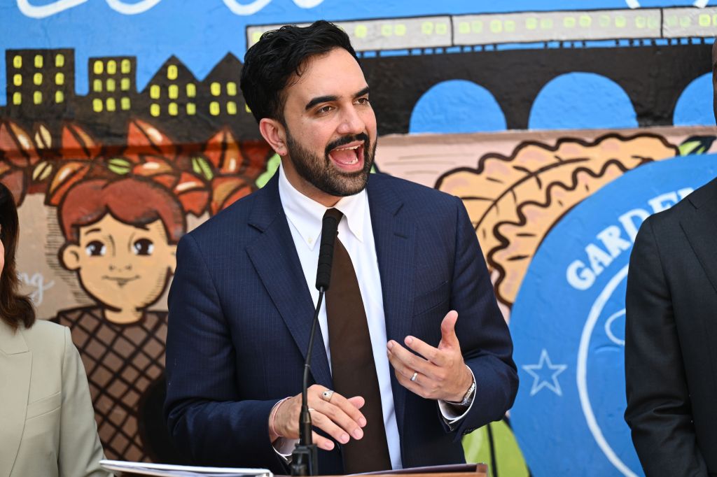Mayor Zohran Mamdani speaks at a press conference, with a colorful mural depicting buildings and children behind him.