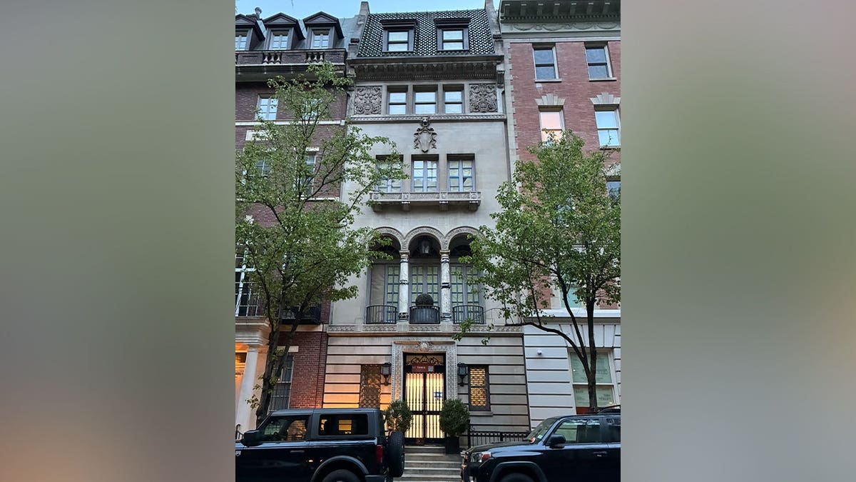 Historic multi-story townhouse facade with ornate stone detailing, arched balcony, and trees lining a city street with parked SUVs.