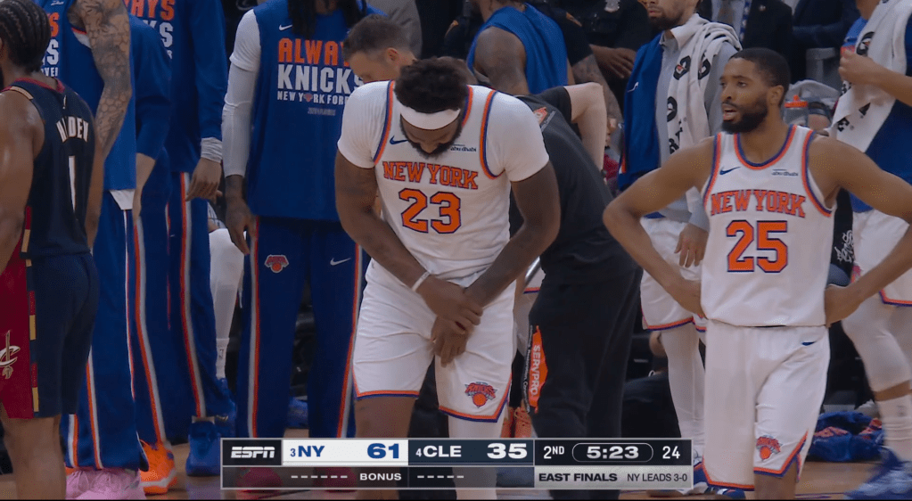 Mitchell Robinson, in a white New York Knicks jersey, clutches his right hand in pain.