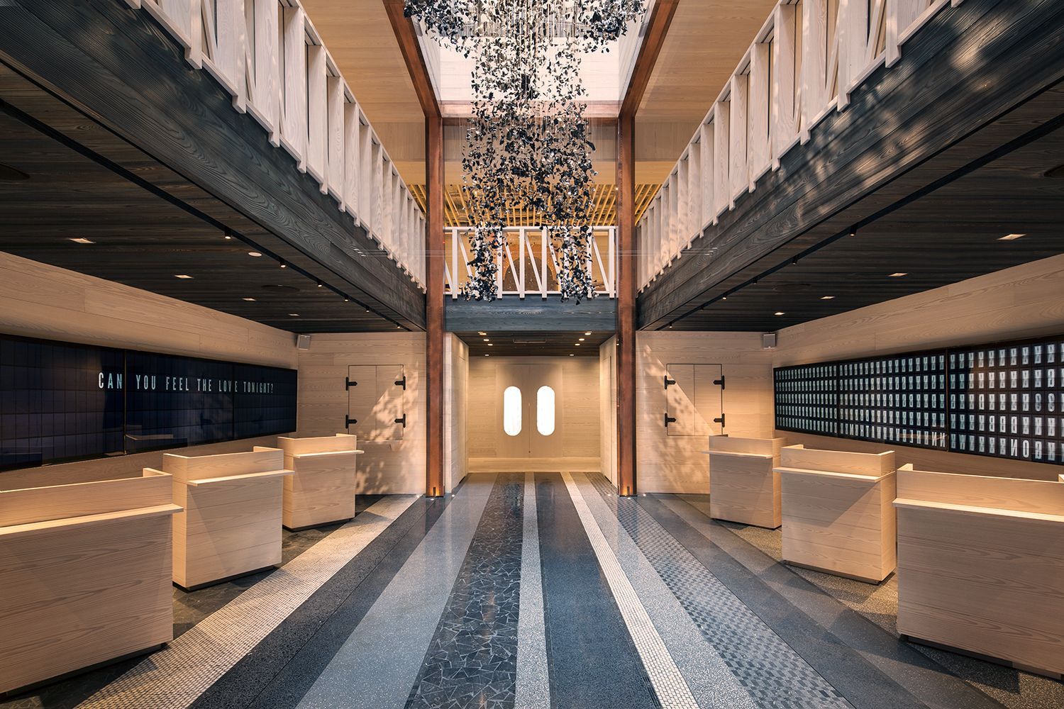 Ground floor check-in area at Moxy Times Square featuring light wood kiosks, a patterned terrazzo floor, and a large hanging bear sculpture suspended in a double-height atrium.