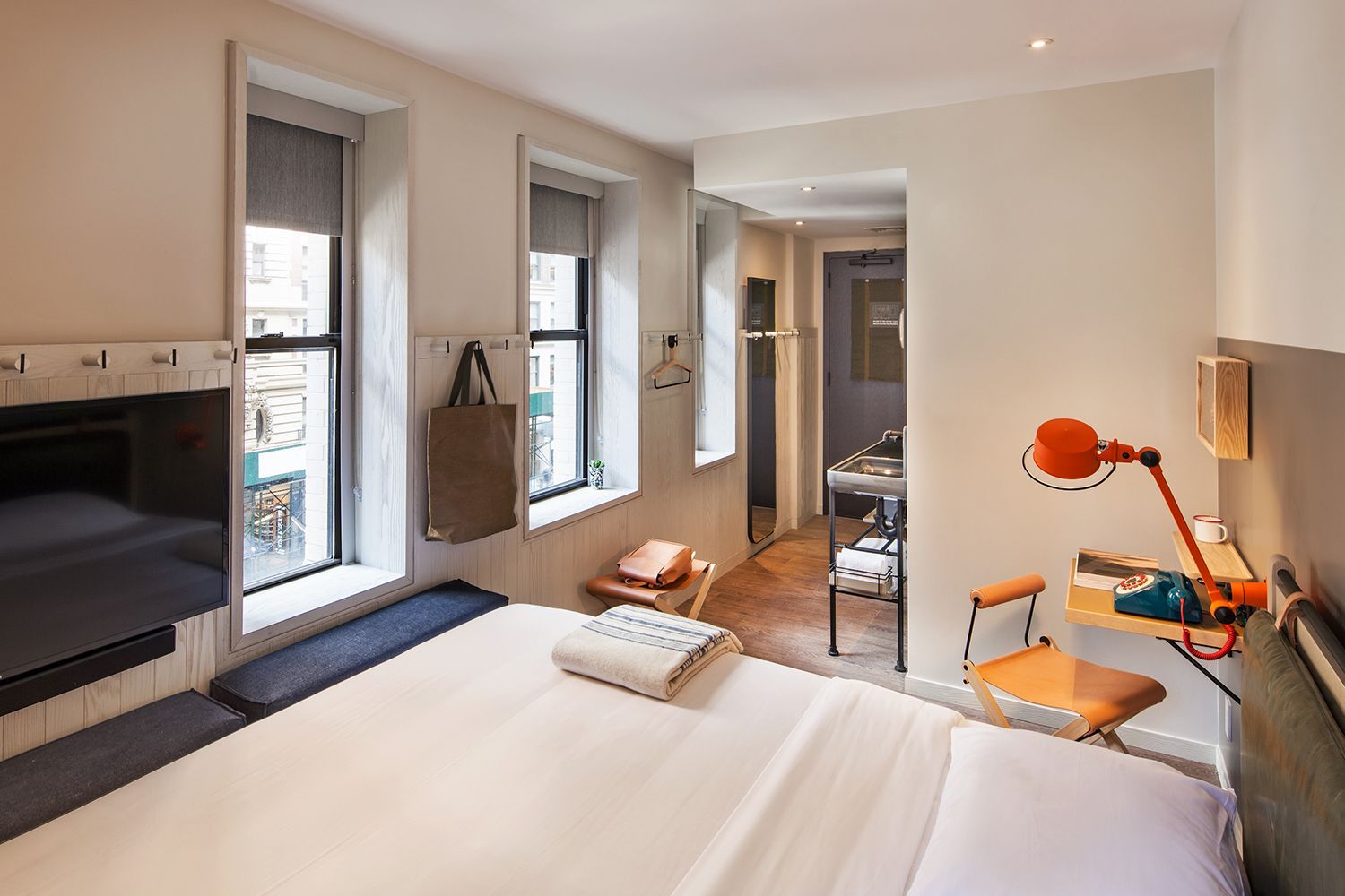 Corner King hotel room at Moxy Times Square showcasing a comfortable bed, large windows with city views, and space-saving foldaway furniture on a pegboard wall.