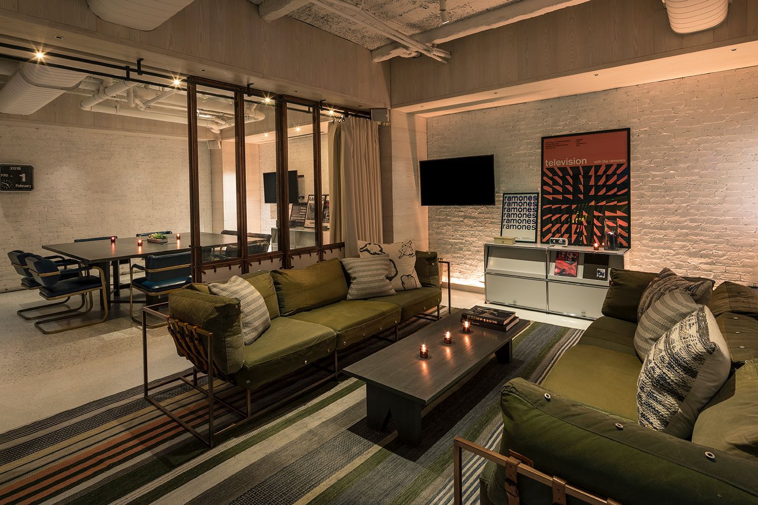 Flexible meeting and lounge space at Moxy Times Square, featuring a green canvas sofa, white brick walls, and glass partitions separating a conference table.