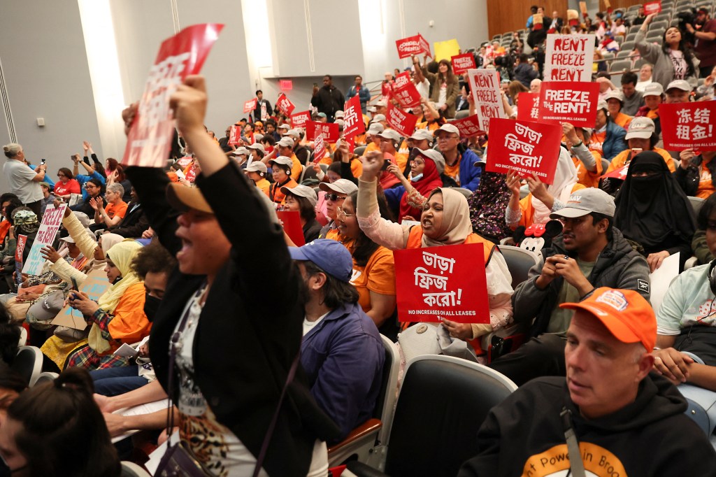 People at a public hearing hold signs that say "Rent Freeze" and "No Increases on All Leases" in multiple languages.