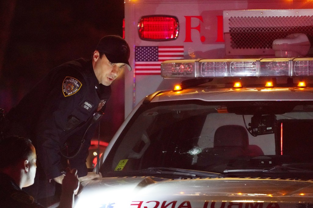 A police officer inspects an ambulance with a bullet hole in its windshield.