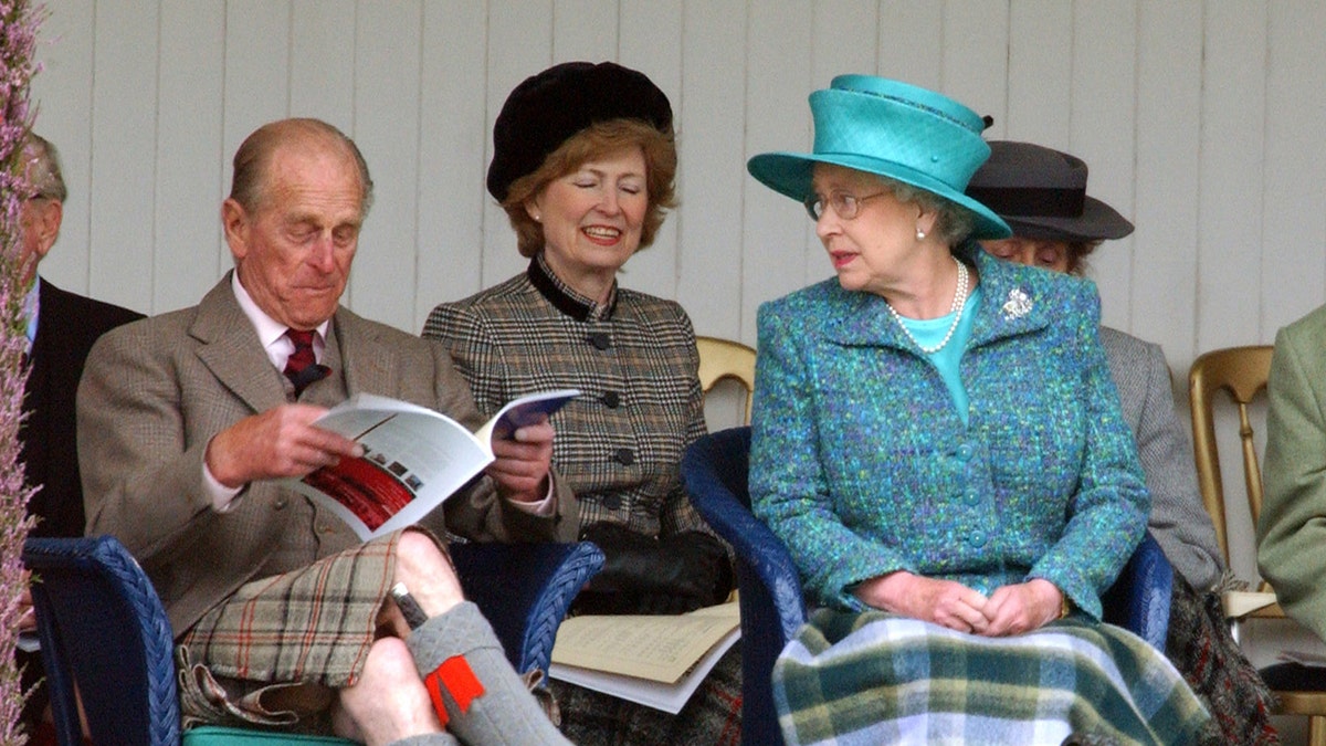 Queen Elizabeth looking at Prince Philip who is reading a book and making funny faces at the Braemar Games