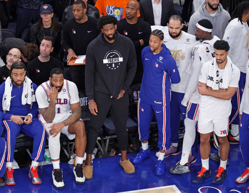 Philadelphia 76ers center Joel Embiid sits on the bench in plain clothes with teammates during a game.
