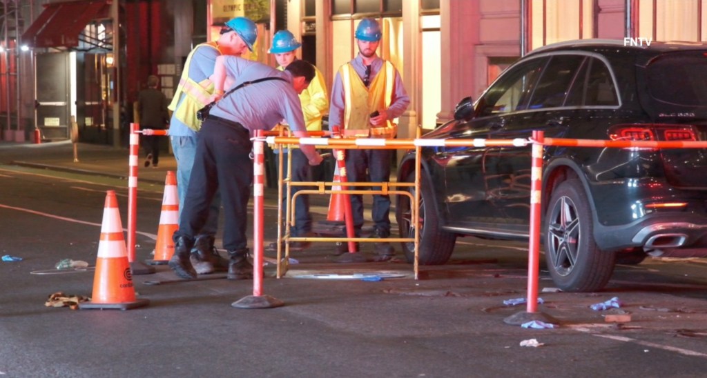 Workers inspect the scene of an accident where a woman fell down an uncovered manhole next to a black Mercedes-Benz SUV.
