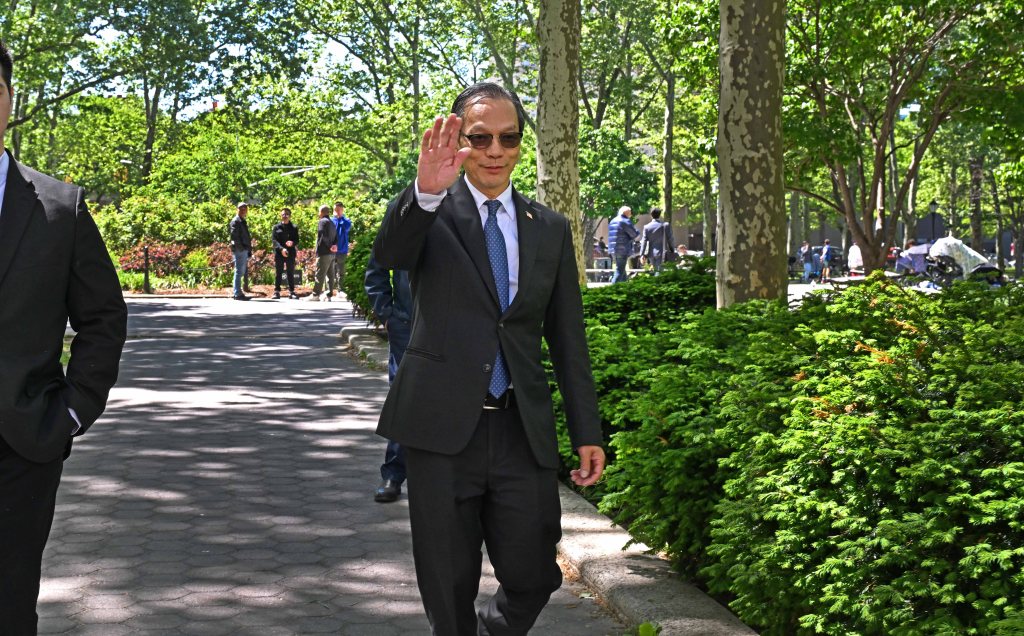 Harry Lu in a suit waves while walking through a park.