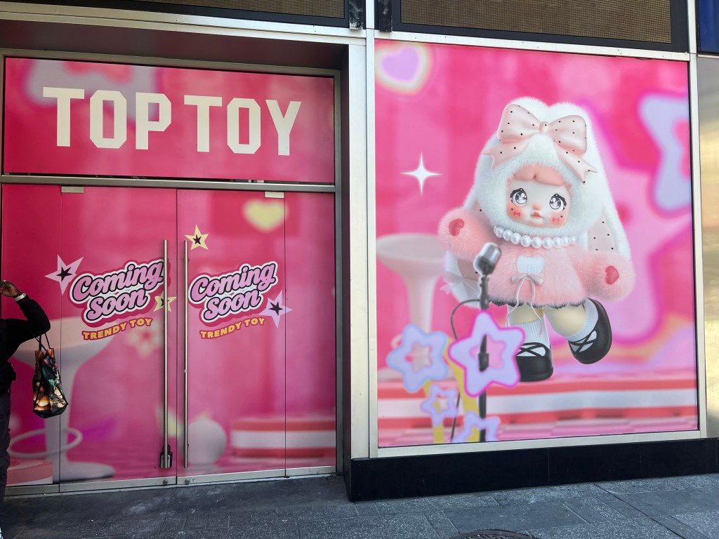 Storefront of the new Top Toy store in Times Square, with pink signs reading "TOP TOY" and "Coming Soon TRENDY TOY" on the left doors, and an illustration of a large cartoon doll in a fluffy pink outfit on the right window.