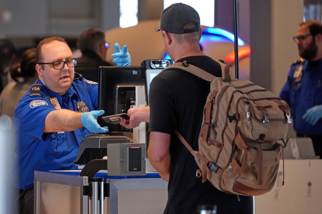 A TSA agent in blue uniform and gloves hands a passport back to a traveler with a backpack.
