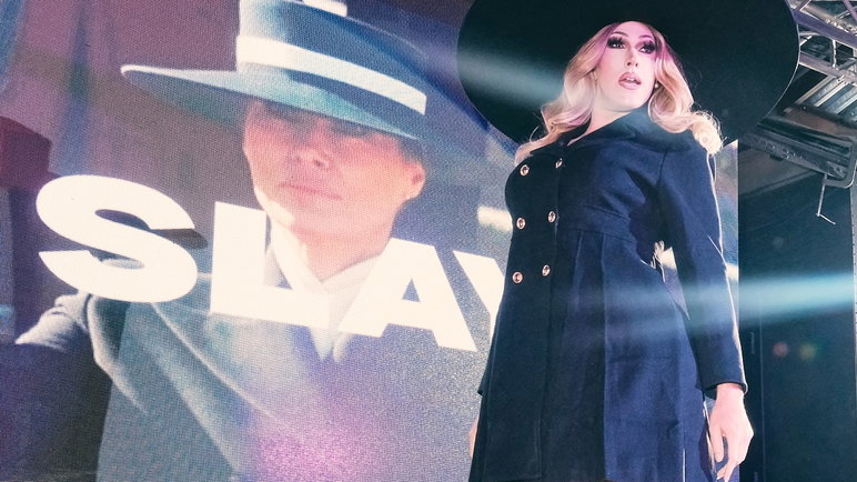 The performer wears a black button-down dress and wide-brimmed hat in front of a screen showing Melania Trump with the word