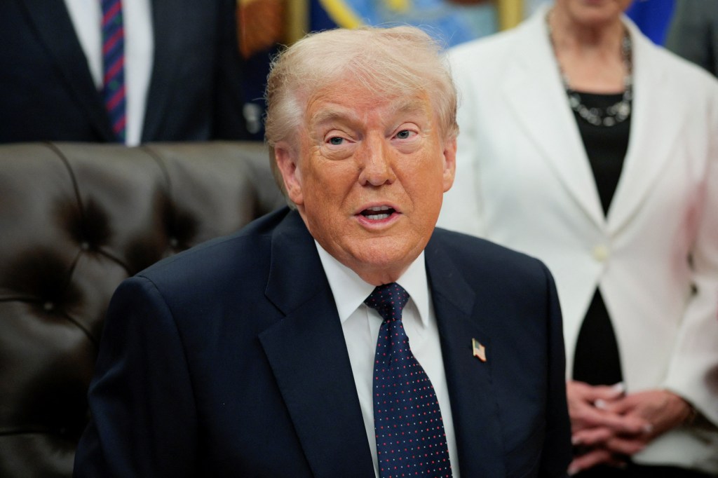 U.S. President Donald Trump in a dark suit and blue tie, speaking at an event in the Oval Office.