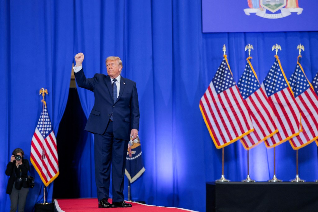 U.S. President Donald Trump stands on a red carpet, raising his right fist, with blue curtains and American flags in the background.