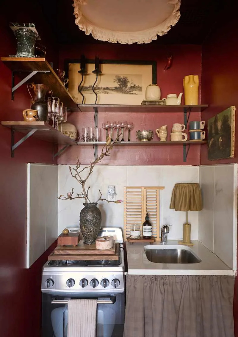Cozy kitchen with deep red walls, vintage decor, a gas stove, and a sink with a wooden shelf and decorative vase.