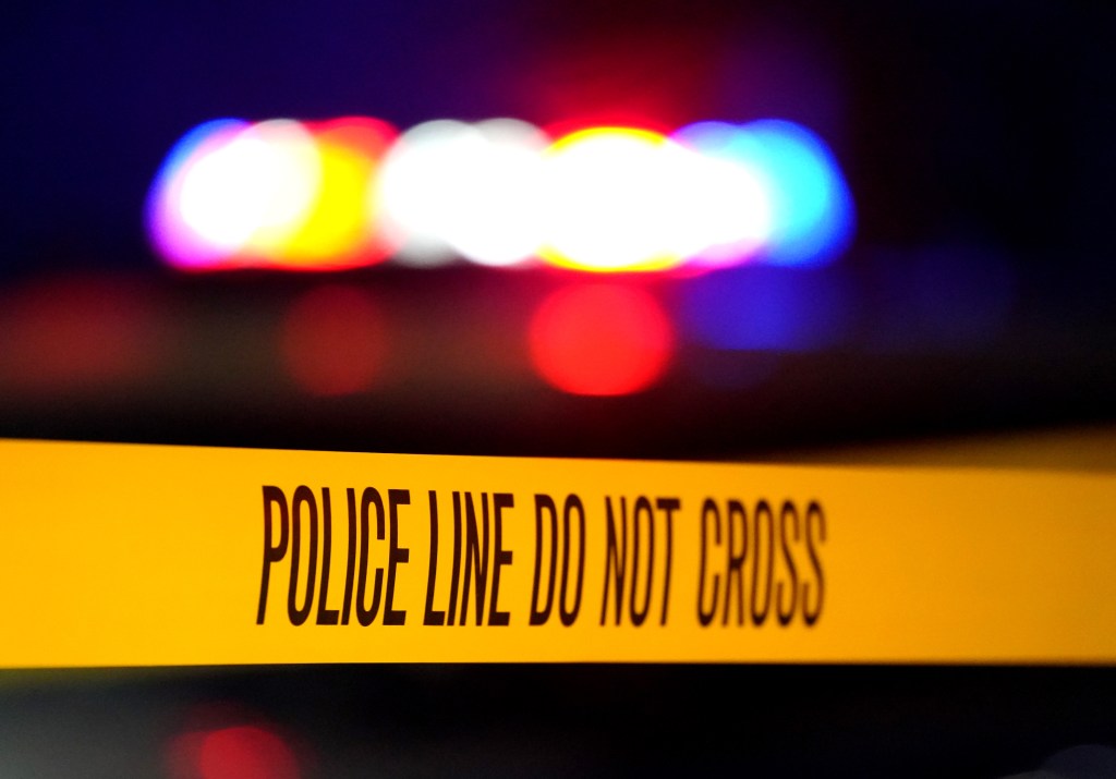 Yellow police tape with the words "POLICE LINE DO NOT CROSS" across it, with blurred red, white, and blue lights in the background.