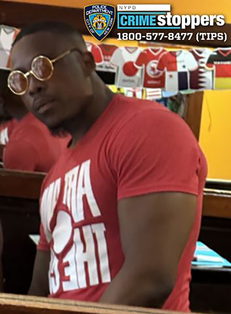 Still image from surveillance video of a dark-skinned male with gold-rimmed circular sunglasses and a red t-shirt with white text, suspected of sexual assault.