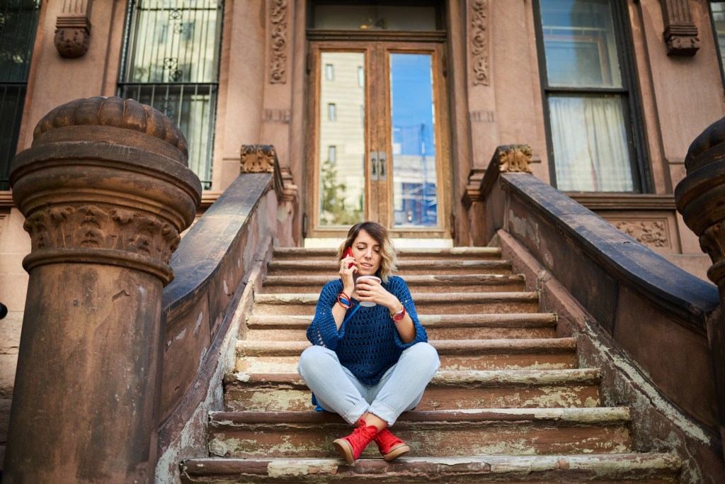A young woman with a red cell phone talking and drinking coffee while sitting on brownstone steps in New York City.