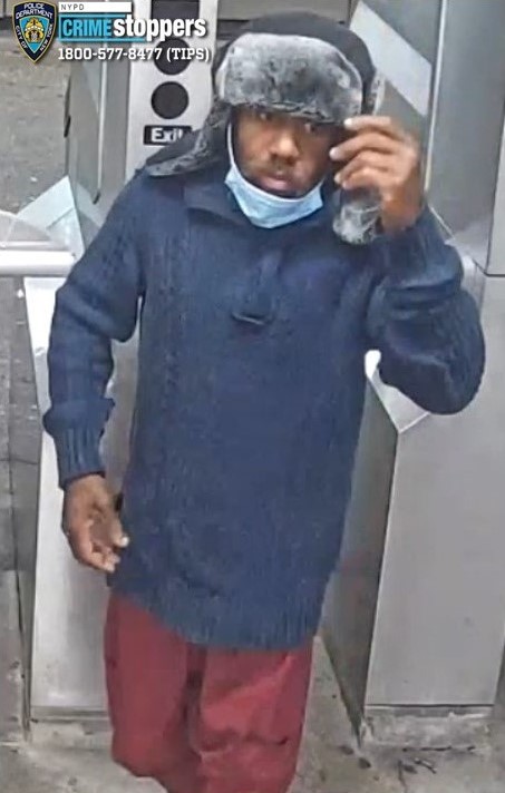 Photo of a male suspect wearing a black and gray fur-lined hat, a blue sweater, a light blue surgical mask pulled down around his chin, and red pants, standing in a subway station.