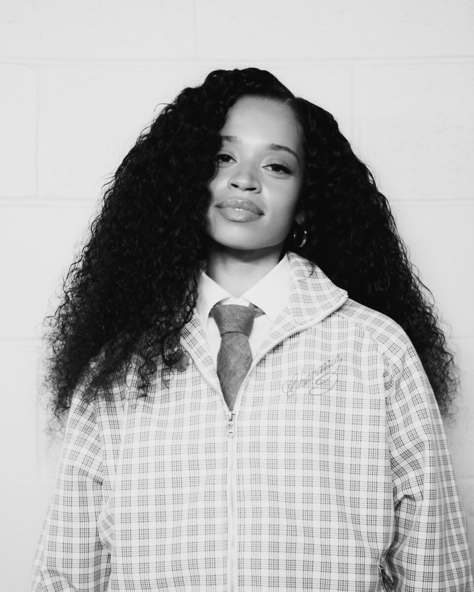 Black-and-white portrait of a woman in a checkered coat and tie standing against a plain wall.