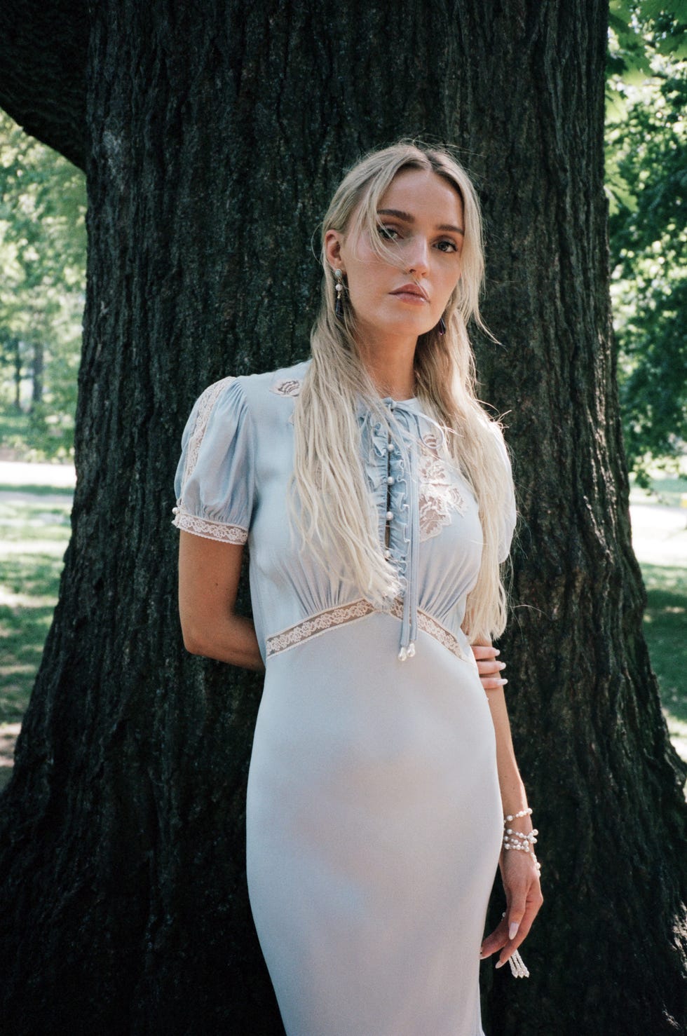 Morgan in a light blue dress stands against a tree in a park.